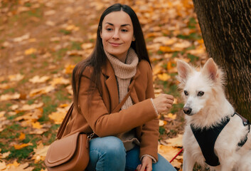A woman with long dark hair, wearing a beige coat and sweater, sits on the ground next to her white...