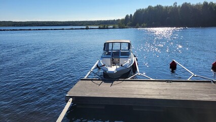 A well-maintained boat sits at a wooden dock beside serene blue waters on a sunny day. Lush trees...