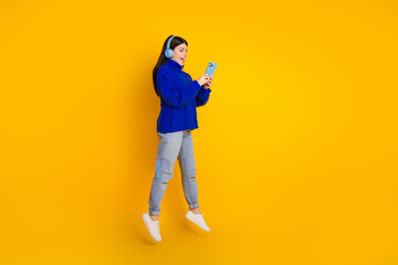 Fototapeta premium Young woman with a blue sweater and headphones, holding a smartphone, surrounded by bright yellow background, showcasing trendy style
