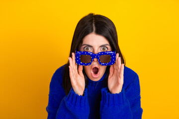 Young woman in blue polka-dot glasses expressing surprise against vibrant yellow background in cozy sweater