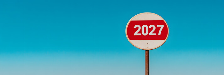Sign against a vibrant blue sky displaying "2027" in bold white numbers on a red band across a white circular sign, mounted on a metal pole. Minimalist future.