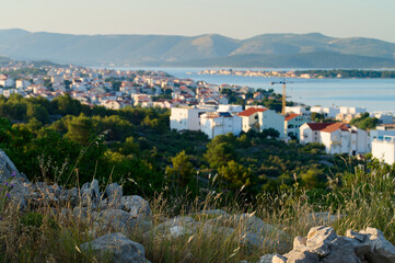 Fototapeta premium Golden sunlight bathes a serene coastal village, nestled among green hills, as the calm sea glimmers. Houses with red roofs create a picturesque landscape filled with tranquility.