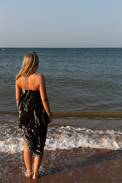 Mujer con vestido moj&aacute;ndose los pies en la orilla del mar.