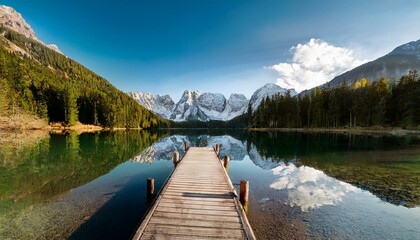 Beautiful wide shot of a lake and mountain in the background and a small deck leading to the lake, with pine trees, clouds, and snow covered mouintain tops 
