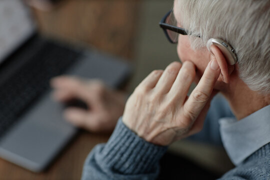 Close up on elderly mans hand tuning up hearing aid behind ear by pressing finger on device controlling volume of sound while working on laptop at home, assistive technology use, copy space