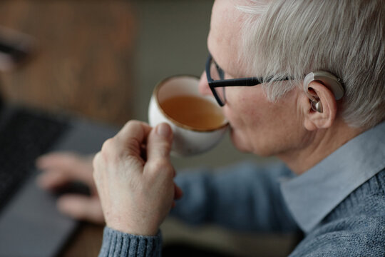 Side view of elderly deafened man wearing hearing aid behind ear using laptop while enjoying warm tea drink at home, copy space