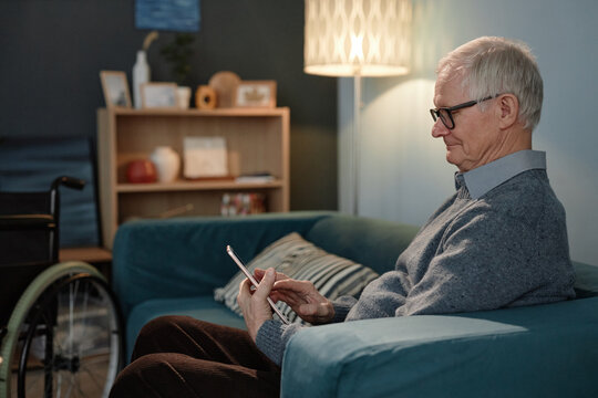 Side view of relaxed senior male wheelchair user typing message on tablet computer wearing glasses while sitting on comfortable couch in cozy living room, copy space - Powered by Adobe