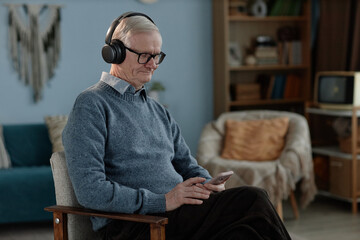 Medium shot of senior tech savvy man listening to podcast in headphones looking at smartphone...