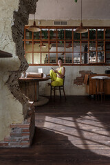 Woman practicing yoga dressed in yellow sits alone in a rustic room with sunlight streaming in through large windows.