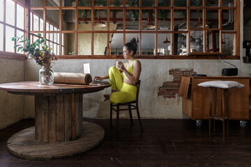 Woman in yellow prepares online yoga class while enjoying a drink in rustic room with wooden floor