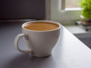 Large white mug full of coffee sitting on a table by a window with a small plant in a pot on the windowsill, enjoying a quiet moment with a warm beverage