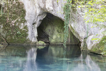 Hidden cave entrance near tranquil blue water surrounded by lush greenery in a serene forest setting