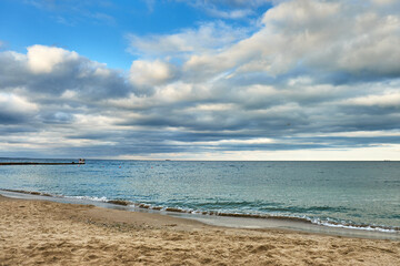 A serene seaside view with soft sand, gentle waves, and a cloudy sky over a calm ocean.