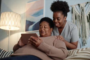 Fototapeta premium Medium shot of African American elderly woman learning how to use online payment services on tablet computer with help of supportive female social worker, while sitting on couch in cozy apartment