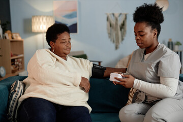 Young African American female nurse using automatic tonometer measuring blood pressure of elderly...