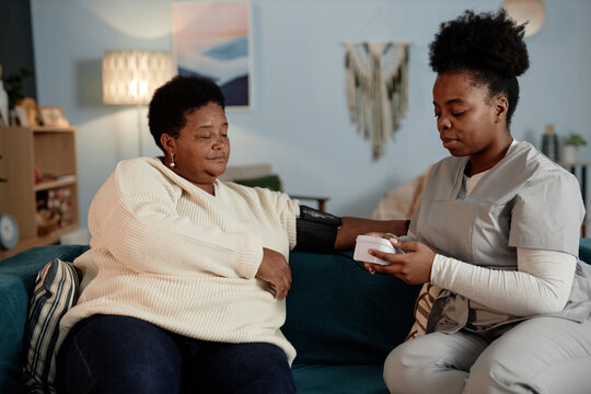 Young African American female nurse using automatic tonometer measuring blood pressure of elderly Black woman during health checkup visit at home - Powered by Adobe