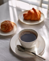 A cozy coffee setup with a cup of coffee, a spoon, and a small plate with a pastry, placed on a clean white tablecloth. Natural light coming from a window creates a relaxing atmosphere.