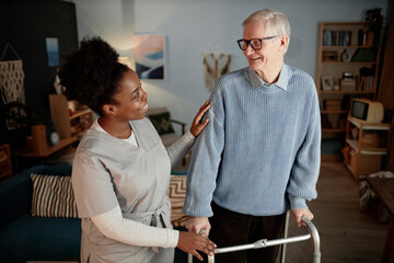 Shot in warm tones of smiling Black female social worker conversing with elderly man walking with frame while helping patient around house in living room, assisted living concept