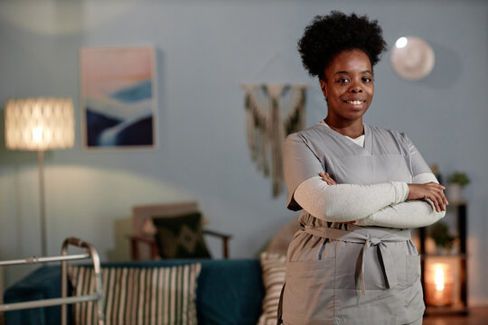 Medium full portrait of African American female social worker wearing grey uniform posing with arms crossed looking at camera with friendly smile during house visit to patient, copy space