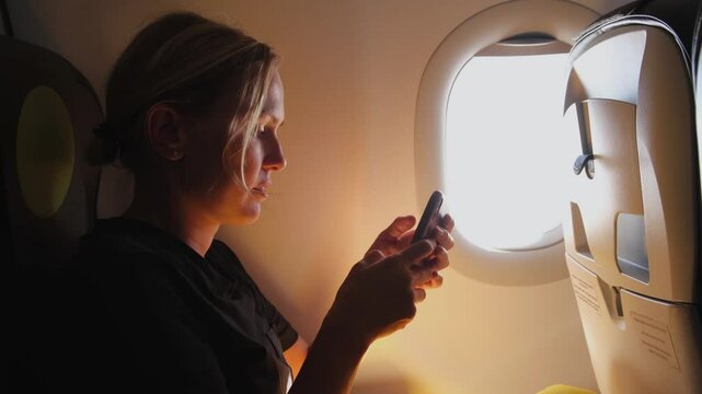 Passenger woman is flying in plane. Girl using phone sitting by airplane window. Traveling female with smartphone inside plane enjoying flight. Woman using wifi connection on board. Traveling girl