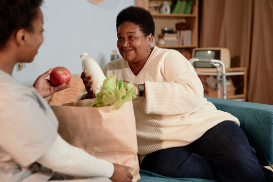 Medium shot in warm tones of happy Black senior woman unpacking paper bag with groceries receiving help from female social worker, delivering food for lonely elderly people at home - Powered by Adobe