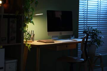 Comfortable workplace with computer and lamp on wooden desk at home office in evening