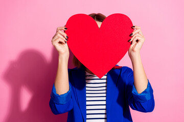 Smiling young woman holding a large red heart shape against a pink background, wearing blue jacket,...