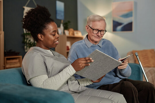 Medium shot of Black female social worker lively discussing pictures in photo album with elderly man, while sitting on couch in living room during home care visit