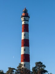 A striking red-and-white Osinovets Lighthouse stands tall against a vibrant blue sky, illuminated by sunlight. The historic maritime beacon, located on the shores of Lake Ladoga, is a significant