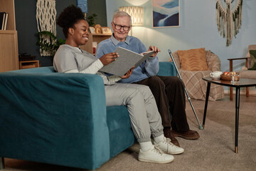 Full length of happy elderly man sitting on couch with female caregiver looking through vintage...