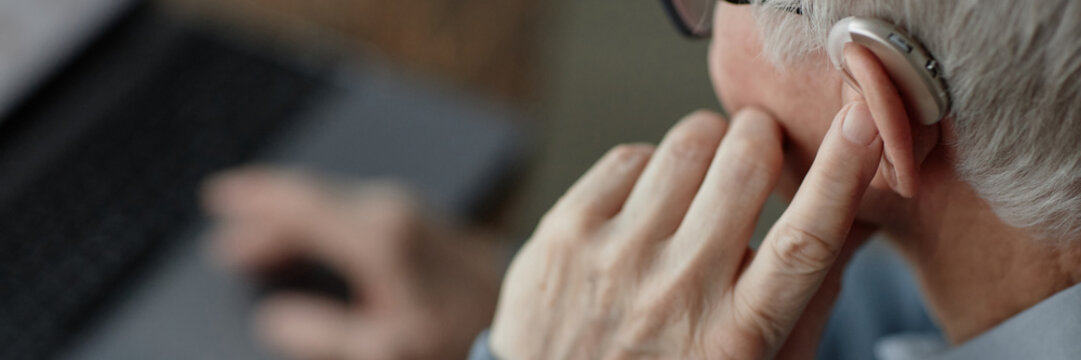 Website header shot of elderly mans hand tuning up hearing aid behind ear by pressing finger on device controlling volume of sound indoors, assistive technology use, copy space