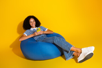 Smiling young woman relaxing on a bean bag in a casual striped outfit using a smartphone against a...
