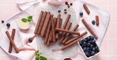 Board with delicious chocolate wafer rolls, blueberries, condensed milk and coffee beans on pink tile background