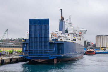 Large ferry boat docked at a port. White and blue ferry boat is moored at pier. Modern ship with ramp is docked at busy port