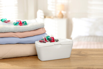 Detergent capsules in container and clean laundry on wooden table indoors. Space for text