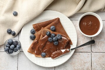 Delicious chocolate crepes with sweet paste and blueberries on light tiled table, flat lay