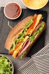 Delicious baguette sandwich with ham and ingredients on grey table, flat lay