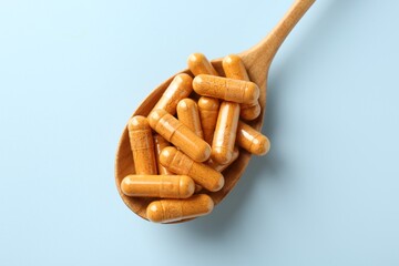 Turmeric capsules in wooden spoon on light blue background, top view