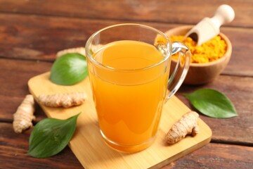 Aromatic turmeric tea in glass mug, roots, powder and green leaves on wooden table, closeup