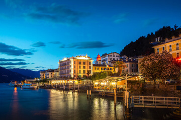 Naklejka premium Evening lights reflect on the tranquil waters of Lake Como, highlighting the charming architecture of Bellagio. The scene captures the vibrant ambiance of Bellagio at night