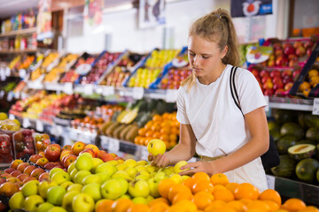 Focused fifteen-year-old girl who came to the store for shopping, chooses ripe apples, standing at the counter
