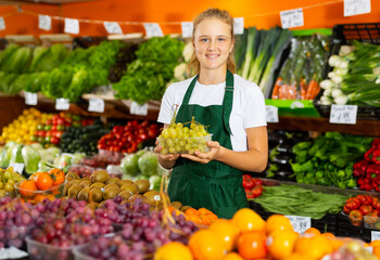 Portrait of a smiling fifteen-year-old girl who works part-time in a store as a trainee seller, standing at the counter, ..holding a box of grapes in her hands