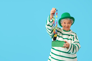 Surprised senior woman in leprechaun hat open gift box for St. Patrick's Day celebration on blue...