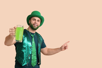 Young man in leprechaun hat with green beard holding glass of beer pointing at something on beige...
