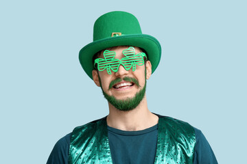 Young man in leprechaun hat and decorative glasses in shape of clover with green beard on blue background. St. Patrick's Day celebration