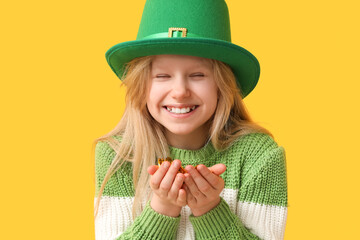 Cute little girl in leprechaun hat with coins on yellow background. St. Patrick's Day celebration