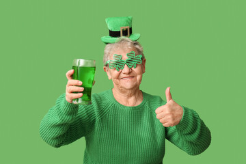 Senior woman in leprechaun hat and decorative glasses with glass of beer showing thumb-up gesture...