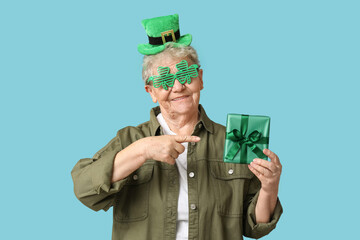Senior woman in leprechaun hat and decorative glasses pointing at gift box on blue background. St....