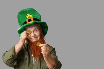 Senior woman in leprechaun hat with red beard holding coins on grey background. St. Patrick's Day...