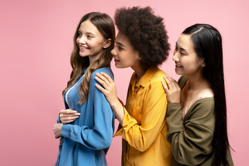 Three smiling young women sharing secrets on pink background